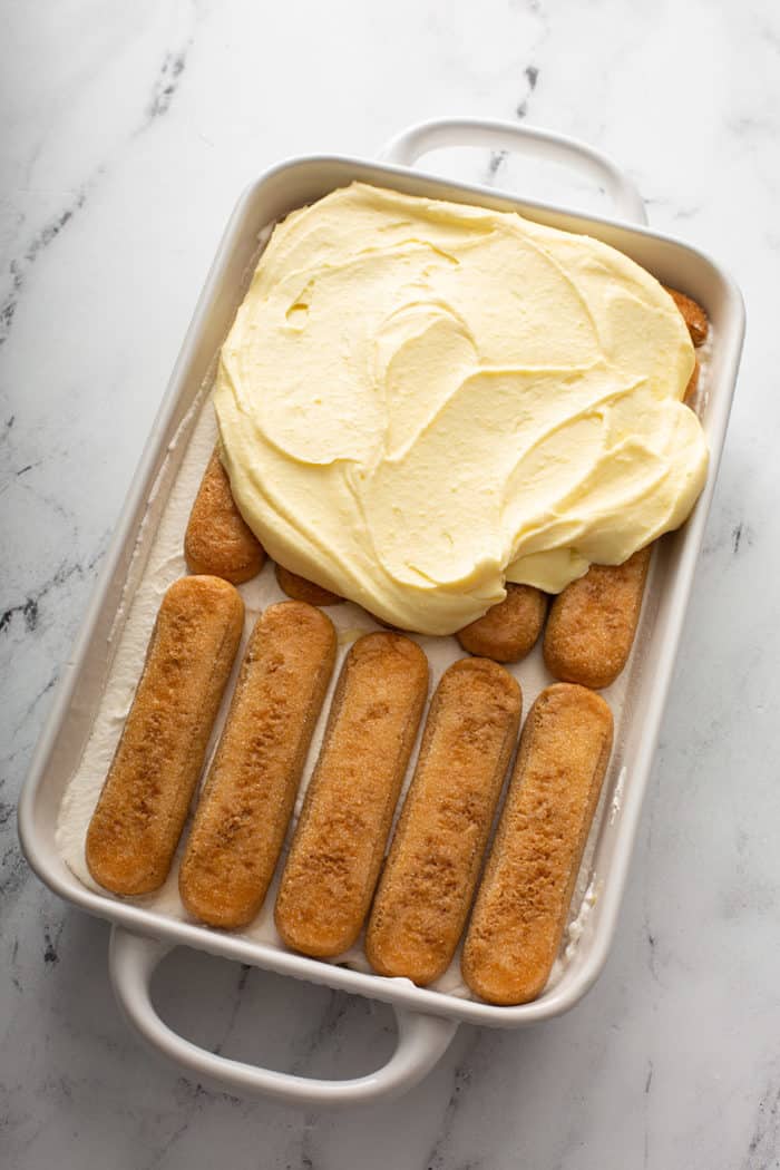 Mascarpone cream being spread on top of soaked ladyfingers in a white baking dish