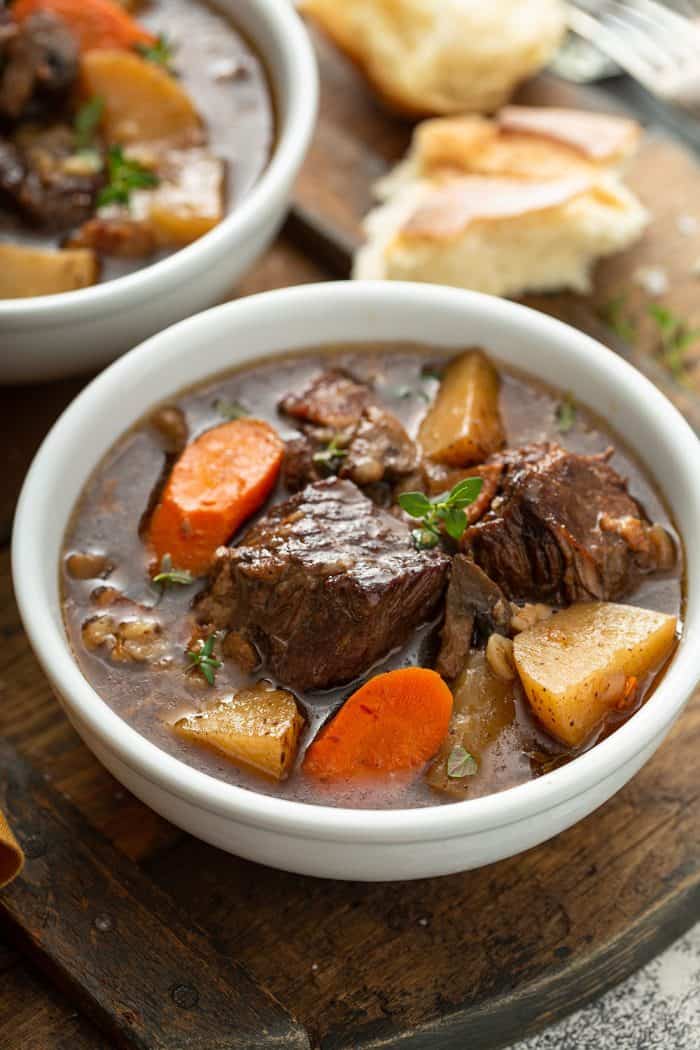 Close up of a beef and barley stew in a white bowl with crusty bread in the background