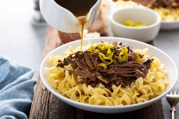 Gravy being poured over Mississippi pot roast on top of egg noodles in a white bowl