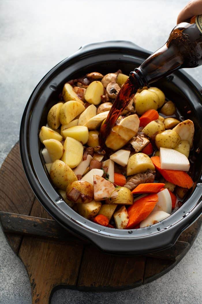 Guinness beer being poured over cut root vegetables in a slow cooker for beef and barley stew