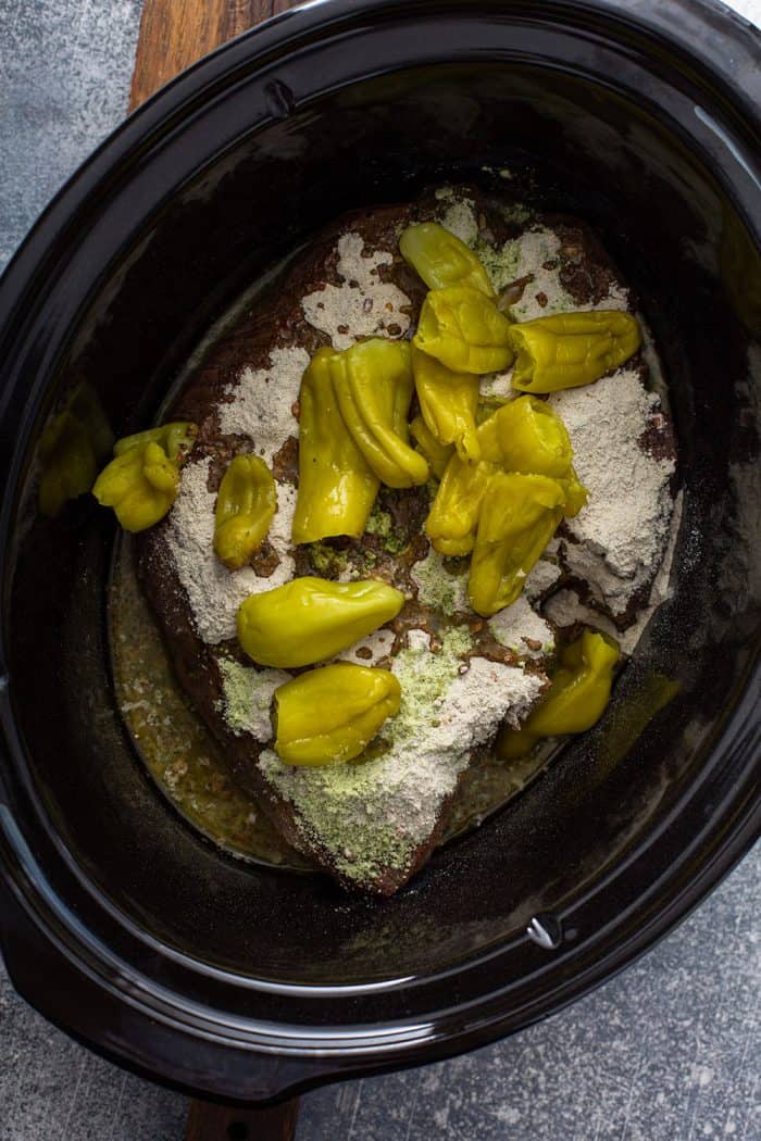Roast topped with seasoning and pepperoncini peppers in a slow cooker crock, ready to cook