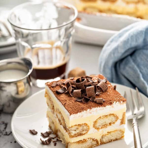 Slice of tiramisu on a white plate with coffee and a baking dish in the background