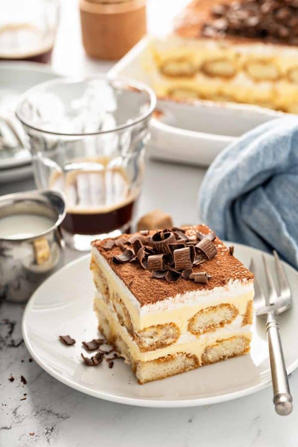 Slice of tiramisu on a white plate with coffee and a baking dish in the background