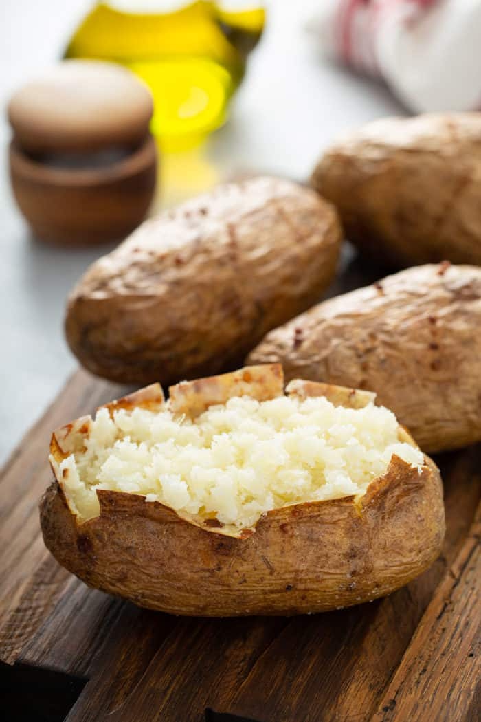 Cut open baked potato on a wooden cutting board with 3 more baked potatoes in the background