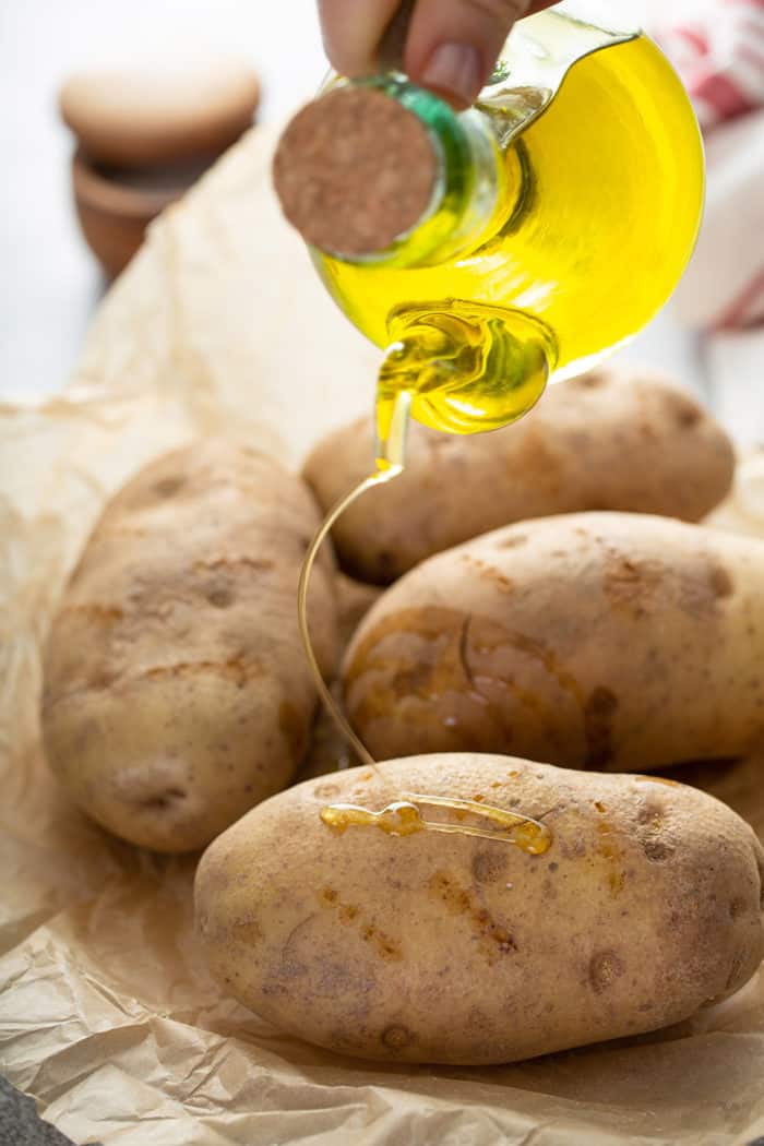 Oil being drizzled over four russet potatoes set on parchment paper