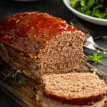 Sliced easy meatloaf with bowls of salad and mashed potatoes in the background