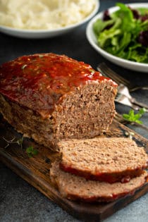 Sliced easy meatloaf with bowls of salad and mashed potatoes in the background