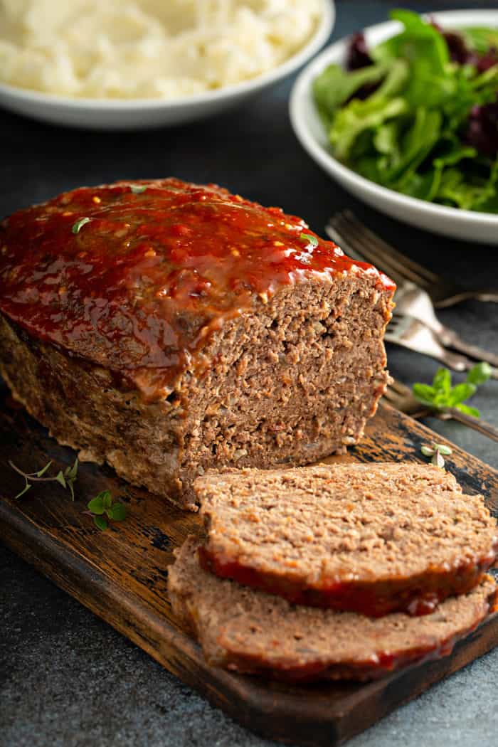 Sliced easy meatloaf with bowls of salad and mashed potatoes in the background