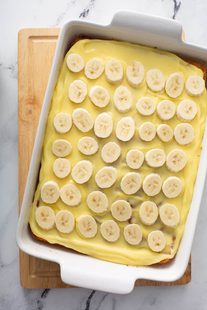 Slices of banana layered on top of pudding-topped banana cake in a white baking dish