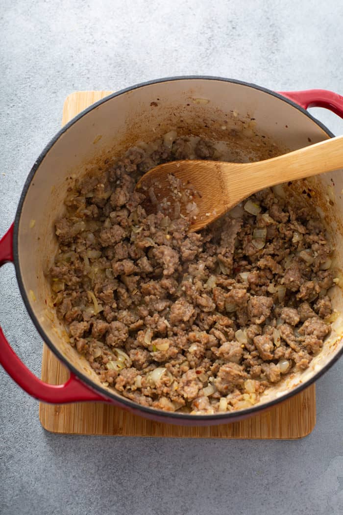 Browned Italian sausage and onions being stirred by a wooden spoon in a large dutch oven