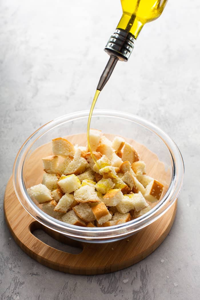 Drizzling olive oil over cubed hamburger buns in a glass bowl