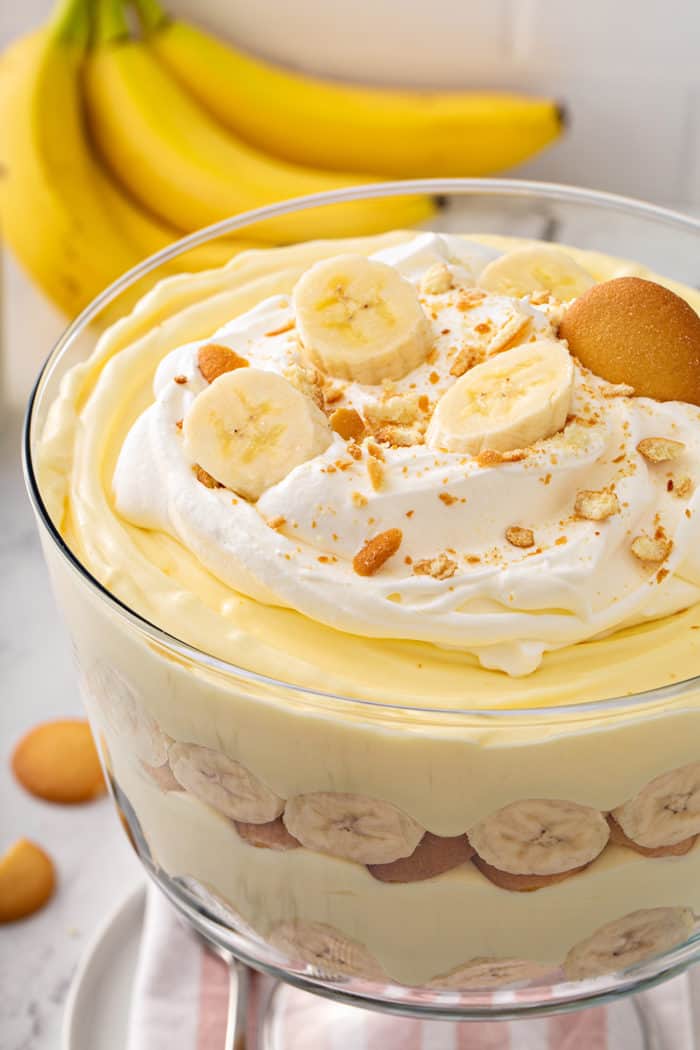 Close up of the top of a layered banana pudding in a trifle dish, topped with whipped cream, bananas and nilla wafers