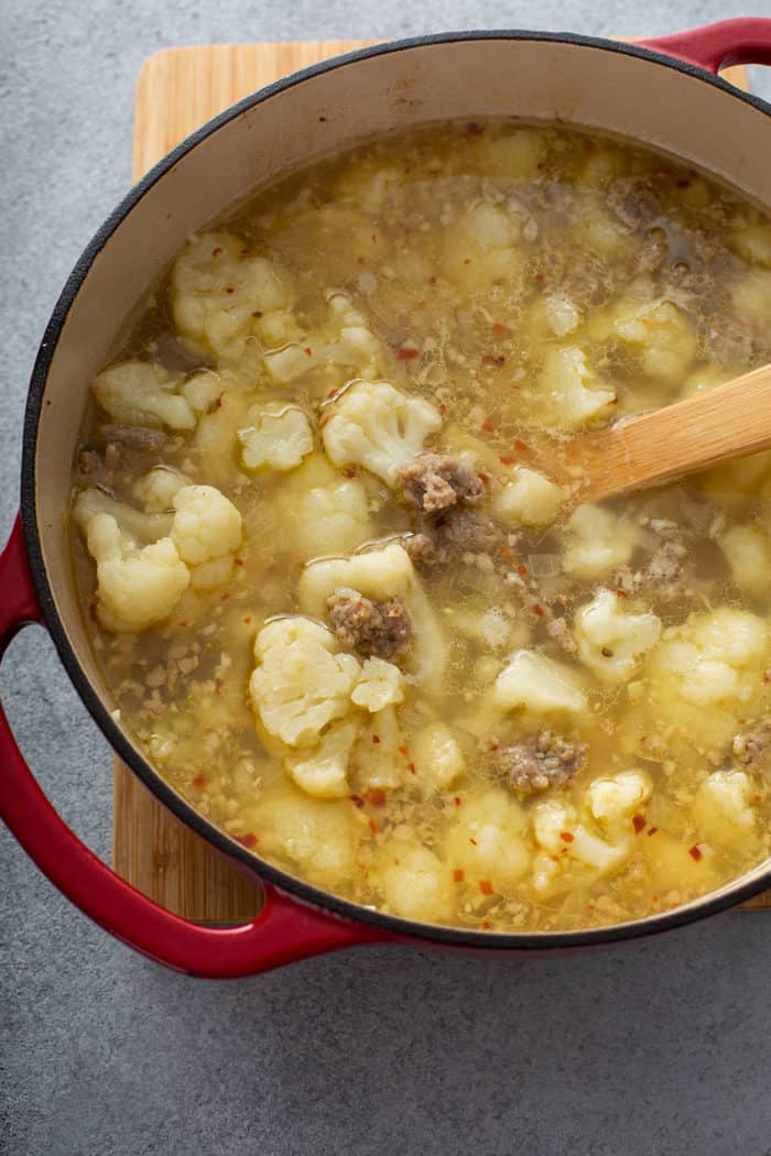 Cauliflower florets and chicken stock being stirred into a pot with cooked Italian sausage for zuppa toscana