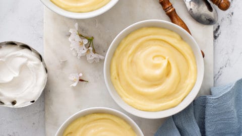 Overhead view of three white bowls of homemade vanilla pudding on a marble board, surrounded by spoons and homemade whipped cream