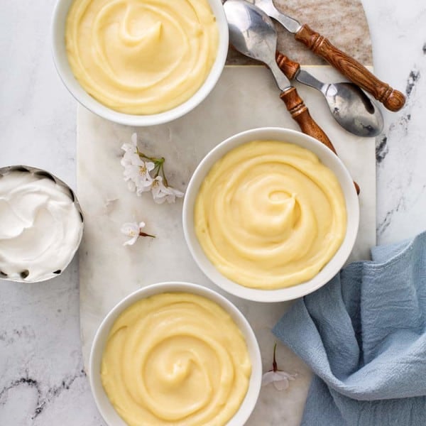 Overhead view of three white bowls of homemade vanilla pudding on a marble board, surrounded by spoons and homemade whipped cream