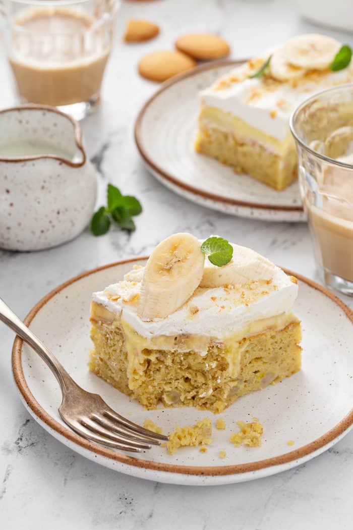 Slice of banana pudding poke cake with a bite taken out of it next to a fork on a white plate, with more plated slices of cake in the background