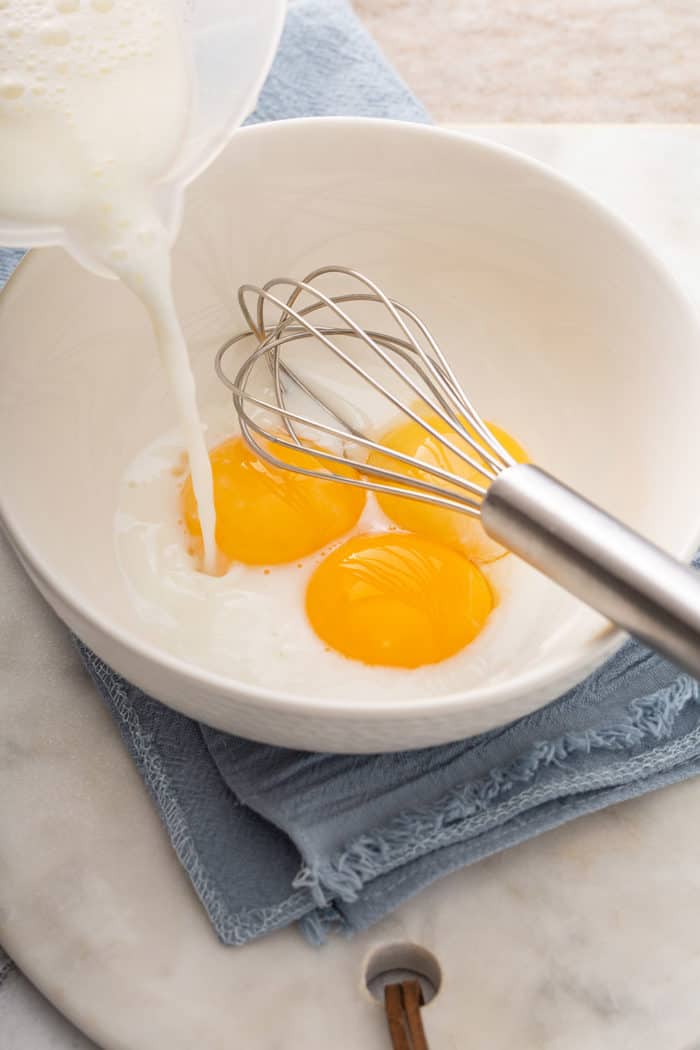 Hot milk being streamed into a white bowl with 3 egg yolks