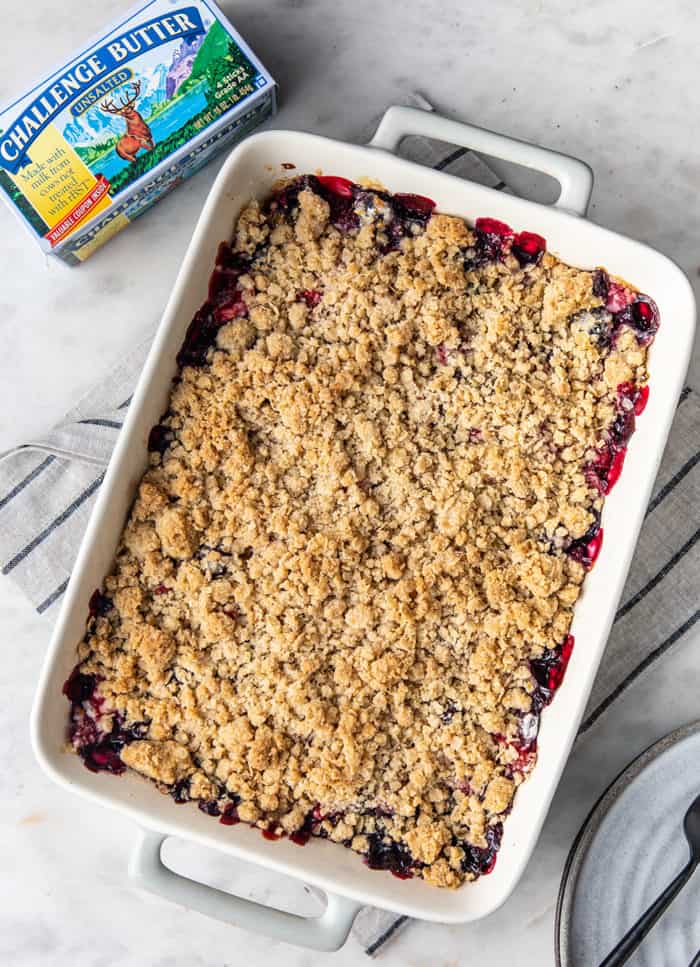 Overhead view of baked berry crisp in a white baking dish, next to a carton of butter