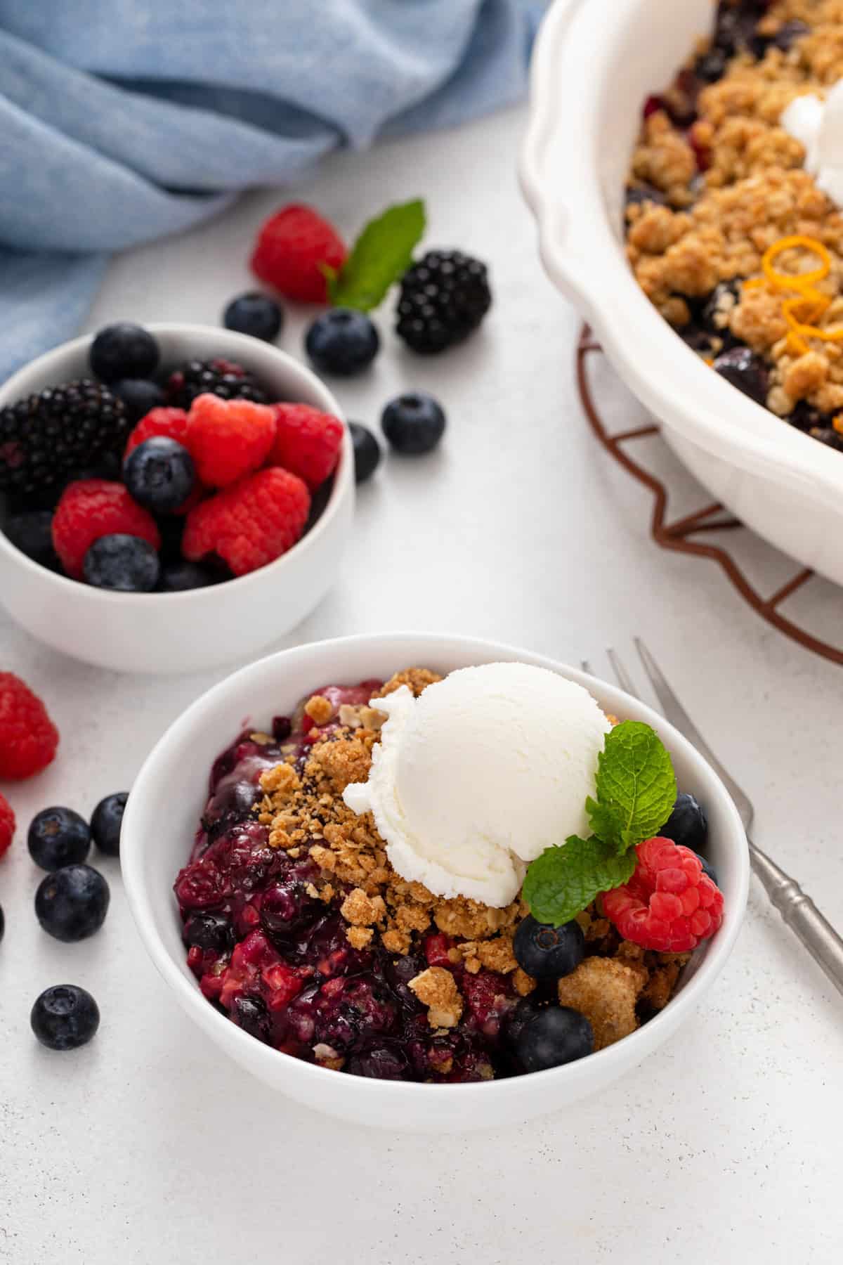 Serving of berry crisp topped with ice cream in a white bowl. A bowl of berries and the rest of the crisp is in the background.