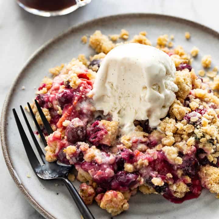 Fork next to berry crisp on a gray plate, with vanilla ice cream starting to melt on top of the crisp