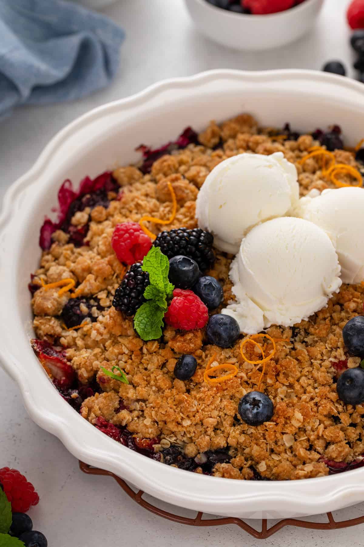 Three scoops of vanilla ice cream and fresh berries on top of a mixed berry crisp in a white baking dish.