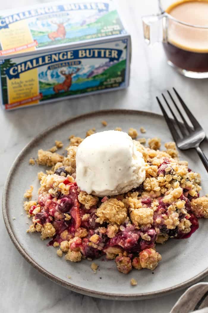 Plate of berry crisp topped with a scoop of vanilla ice cream, with a container of butter in the background