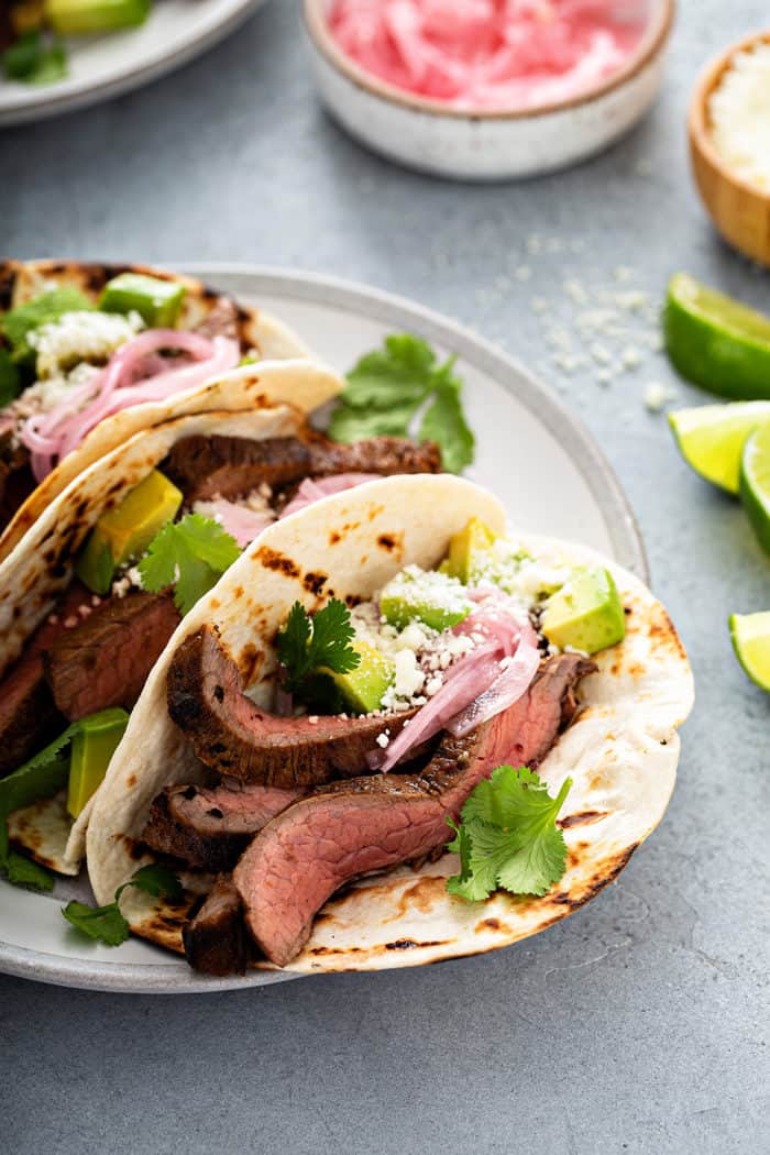 Close up of a flank steak taco in a flour tortilla, topped with avocado, pickled onion and cilantro