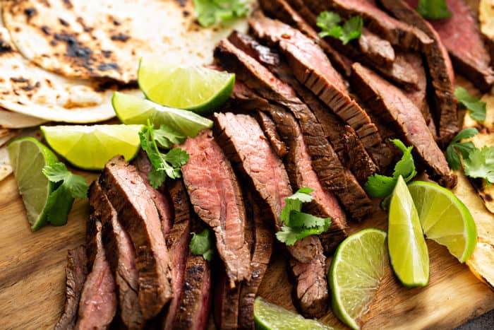 Close up of sliced flank steak surrounded by lime wedges on a wooden board