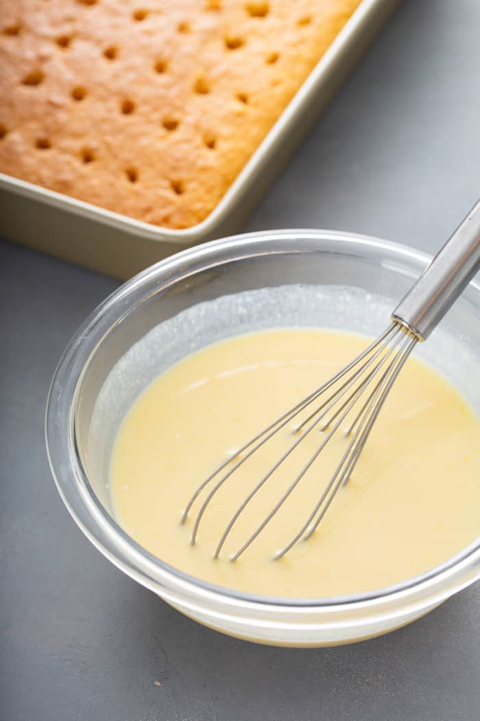 Whisk mixing together coconut pudding in a glass bowl with a baked yellow cake in the background