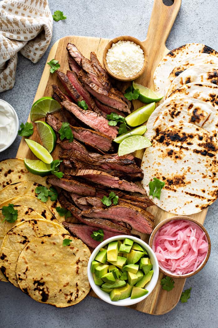 Wooden board with sliced flank steak, charred corn and flour tortillas, bowls of avocado, cotija cheese, and pickled onions, and lime wedges