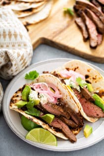 Two flank steak tacos in charred flour tortillas on a white plate with a wooden cutting board in the background