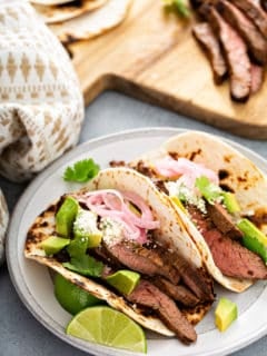Two flank steak tacos in charred flour tortillas on a white plate with a wooden cutting board in the background