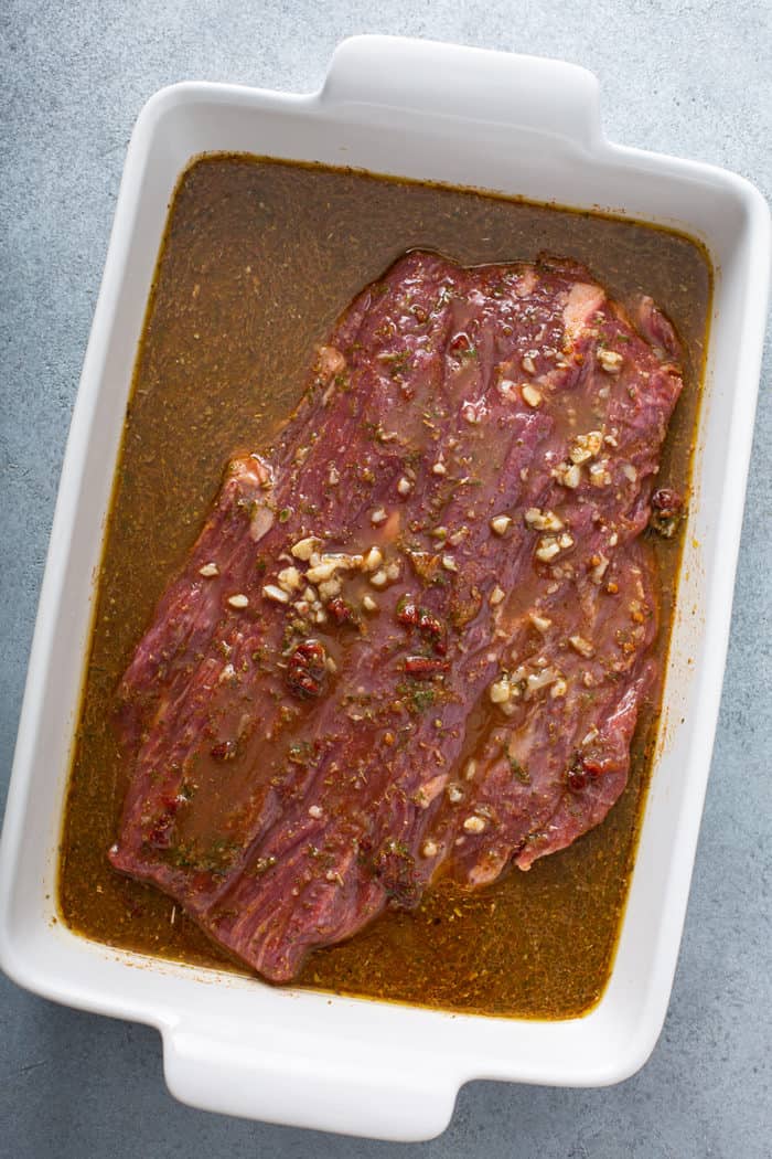 Flank steak covered in marinade in a white baking dish
