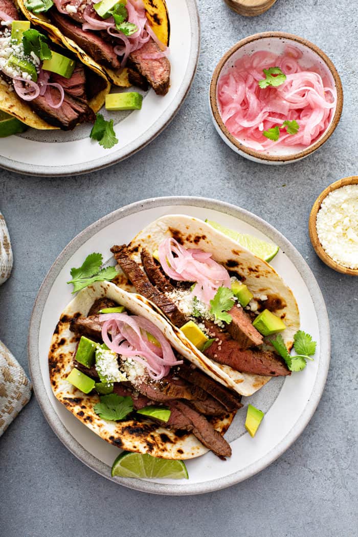 Overhead view of two white plates, each holding multiple flank steak tacos, next to bowls of pickled onions