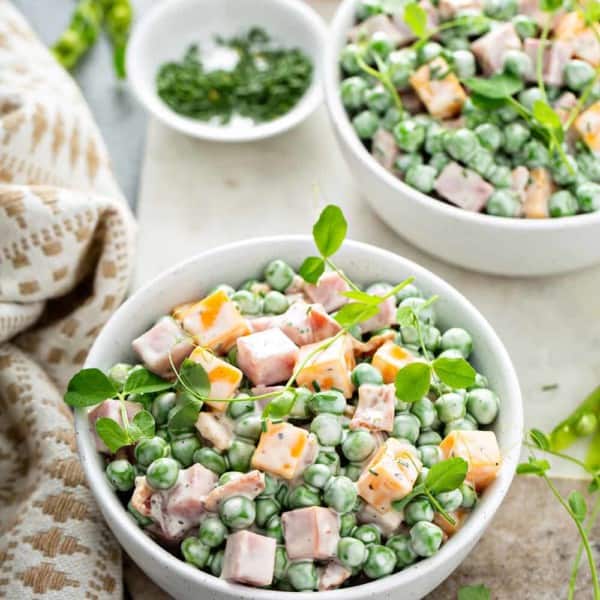 Two bowls filled with Amish pea salad surrounded by fresh herbs