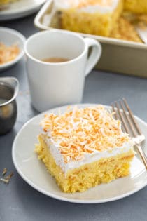 Slice of coconut poke cake next to a fork on a white plate, with a cup of coffee in the background