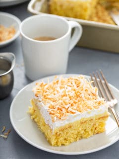 Slice of coconut poke cake next to a fork on a white plate, with a cup of coffee in the background