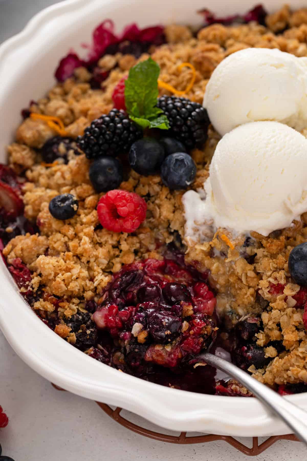 Serving spoon in a white baking dish filled with berry crisp topped with vanilla ice cream.