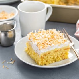 Plated slice of coconut poke cake next to a fork with a pan of cake and cup of coffee in the background