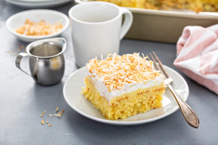 Plated slice of coconut poke cake next to a fork with a pan of cake and cup of coffee in the background