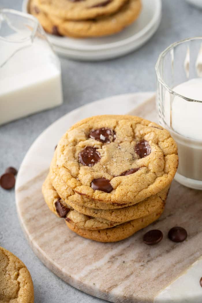 Five brown butter chocolate chip cookies stacked on a wooden board next to a glass of milk