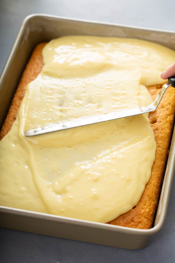 Offset spatula spreading coconut pudding over the top of a yellow cake with holes poked in it