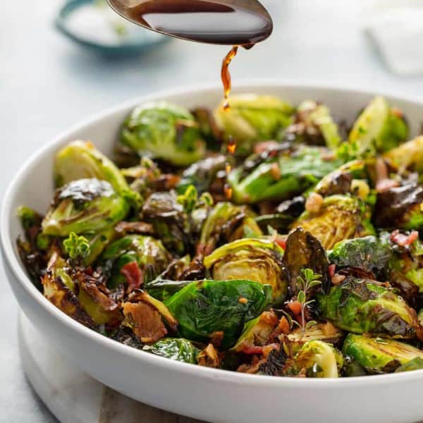 Spoon drizzling balsamic vinegar over air fried brussels sprouts in a white serving bowl