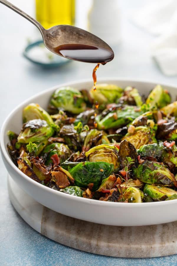 Spoon drizzling balsamic vinegar over air fried brussels sprouts in a white serving bowl