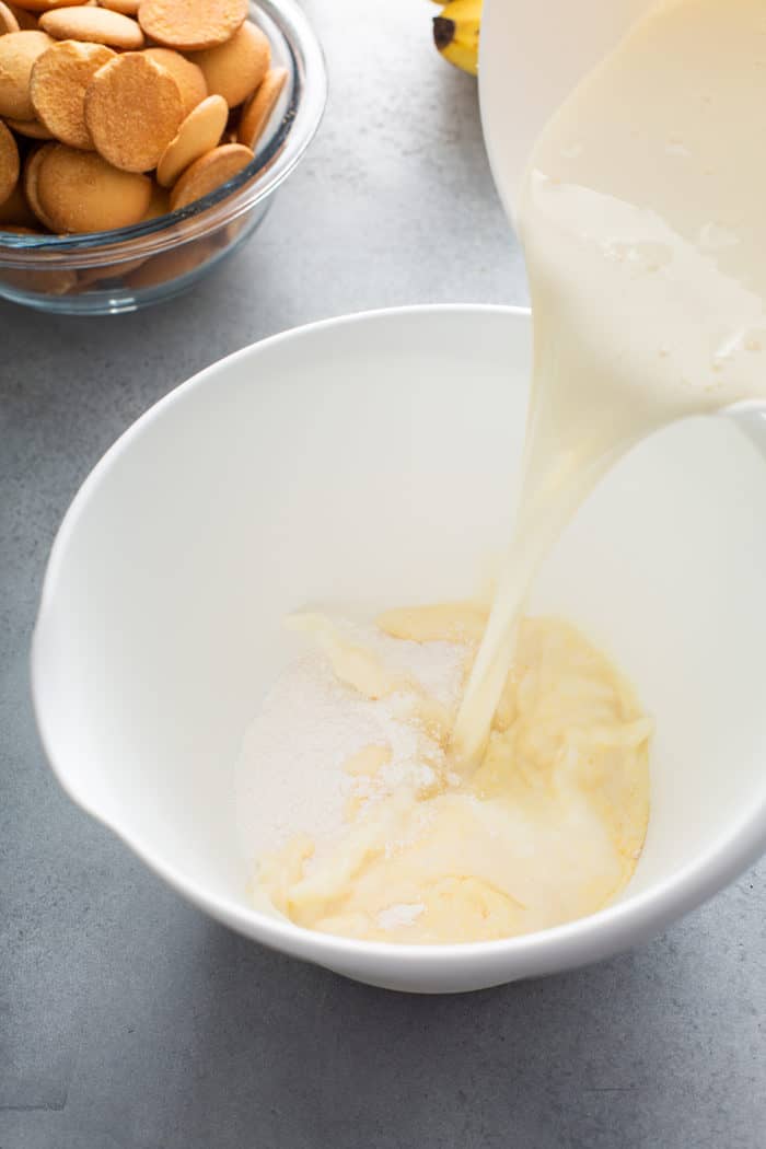 Condensed milk and water mixture being added to instant vanilla pudding mix in a white mixing bowl