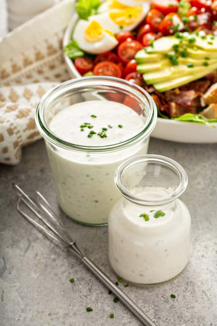 Jar and glass pitcher of homemade ranch dressing on a gray countertop next to a bowl of salad