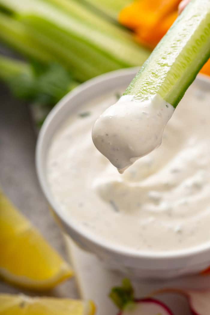 Cucumber stick dipped in ranch dip with a bowl of the dip in the background