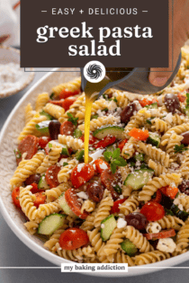 Salad dressing being drizzled over greek pasta salad in a large serving bowl. Text overlay includes recipe name.