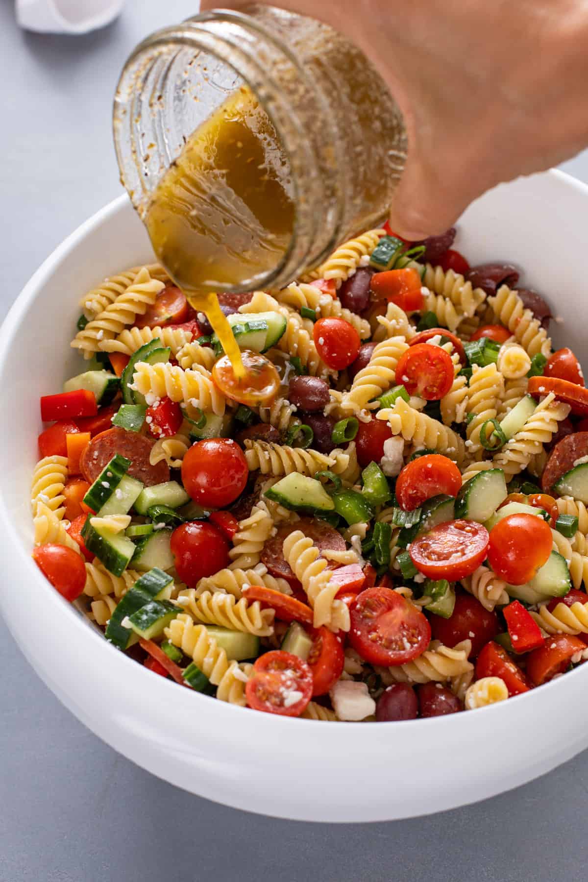Greek salad dressing being poured over pasta salad in a large white mixing bowl.