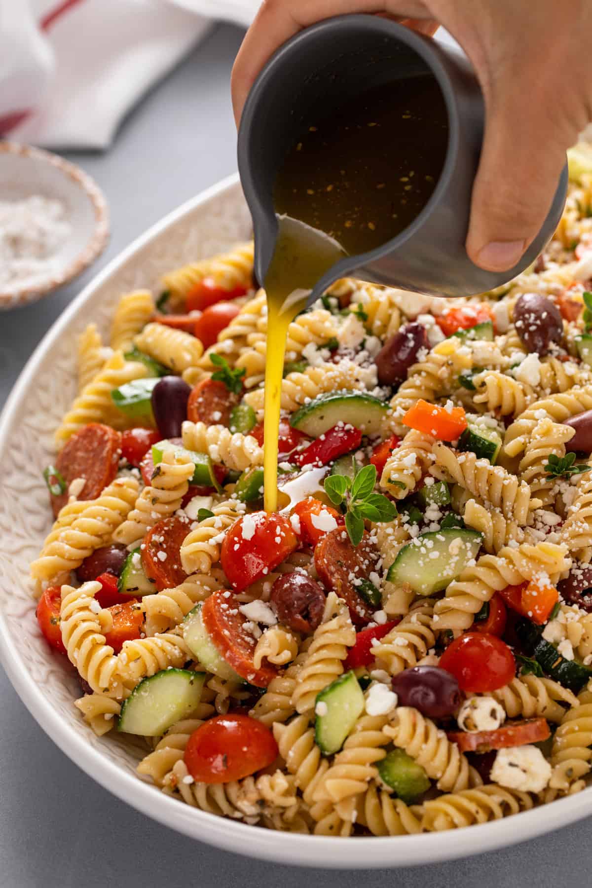 Salad dressing being drizzled over greek pasta salad in a large serving bowl.